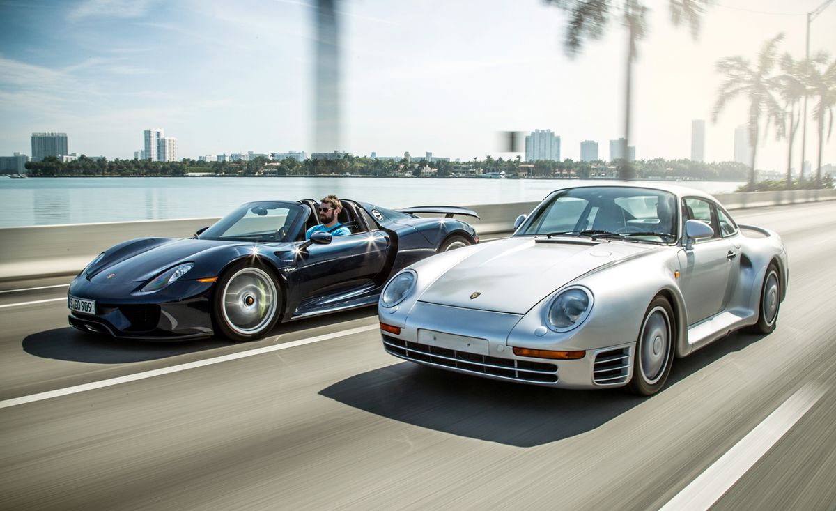 Rolling shot showing the front-angled view of a black Porsche 918 Spyder and silver Porsche 959 on a highway.